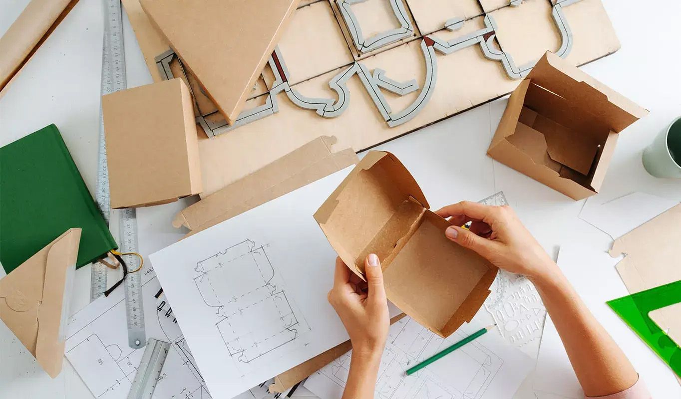 Hände montieren Kartonverpackungen mit Designplänen und Werkzeugen auf einem Tisch.
